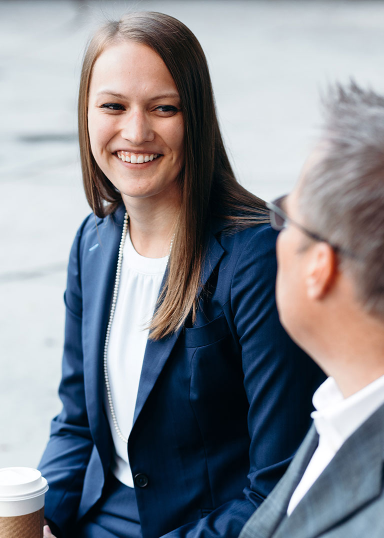 Image of a business man and business woman sitting outside. The woman is in frame and smiling at the man.