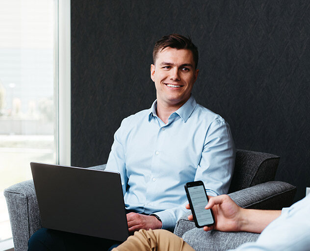 Two men sitting in chairs. One has a phone in his hand, and the other has a laptop in his lap. They are both smiling.
