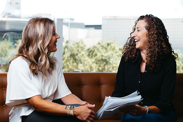Image of two women laughing with each other while sitting on a couch and holding papers